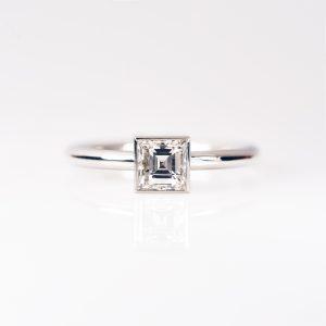 Front view of carre-cut diamond solitaire ring set in polished platinum, against a soft pale grey background.