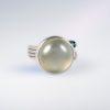 Platinum ring with a wide, ribbon-wrapped band, featuring a large cabochon moonstone, an orange pear-shaped rose-cut diamond, and a small green cabochon tourmaline.